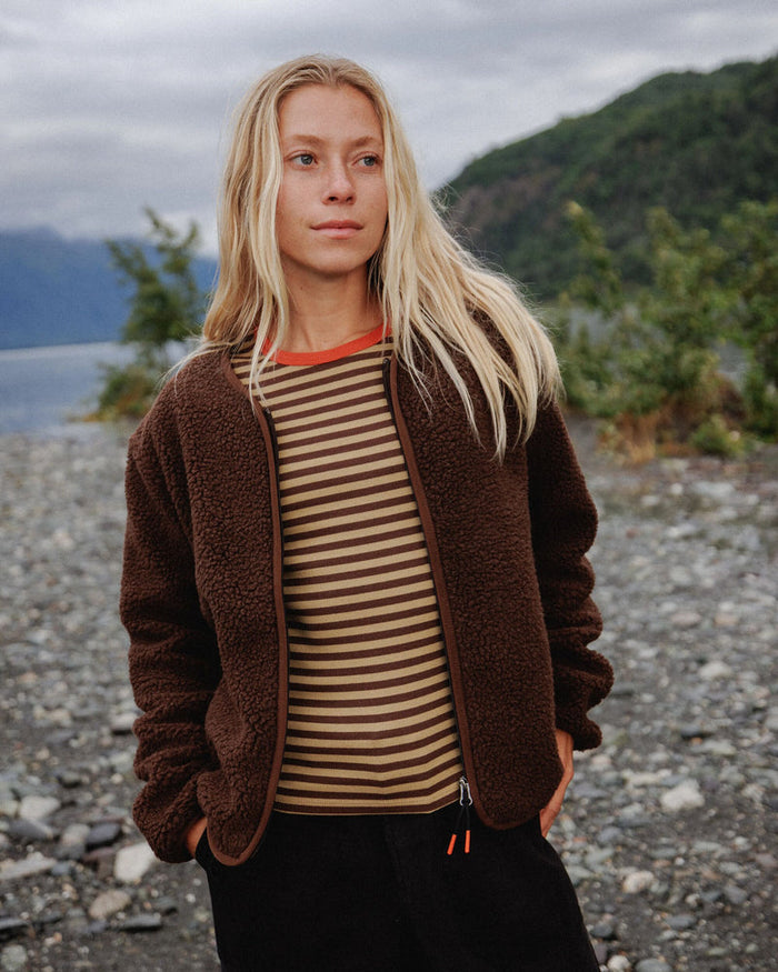 A woman standing outdoors on a rocky lakeshore wearing a brown sherpa zip-front jacket over a beige and brown horizontally striped shirt, with dark pants and a cloudy mountain landscape in the background.