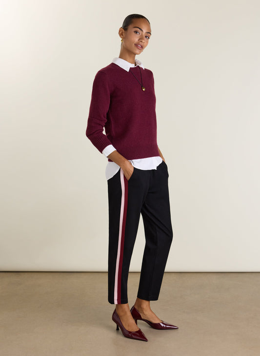A woman modeling a burgundy knit polo sweater with a white collar, paired with black trousers featuring a pink and white side stripe, and burgundy patent leather heels, against a neutral studio background.