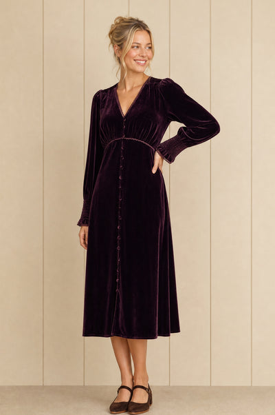 A woman modeling a long-sleeved, button-front velvet midi dress in deep plum purple, standing against a beige paneled wall.