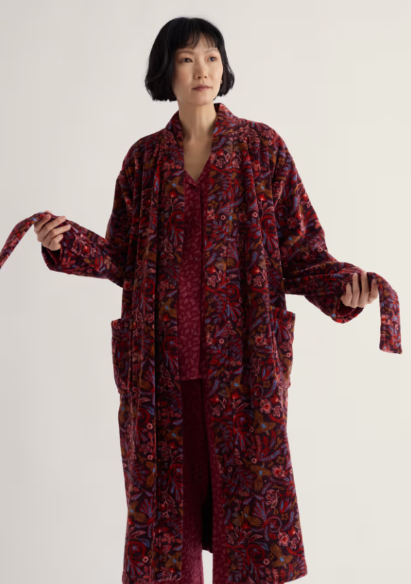A model wearing a long-sleeved, open-front robe with a red, purple, and black floral paisley pattern, paired with matching burgundy pajamas, against a plain off-white studio background.