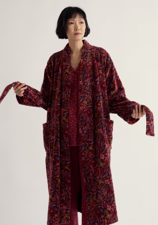 A model wearing a long-sleeved, open-front robe with a red, purple, and black floral paisley pattern, paired with matching burgundy pajamas, against a plain off-white studio background.