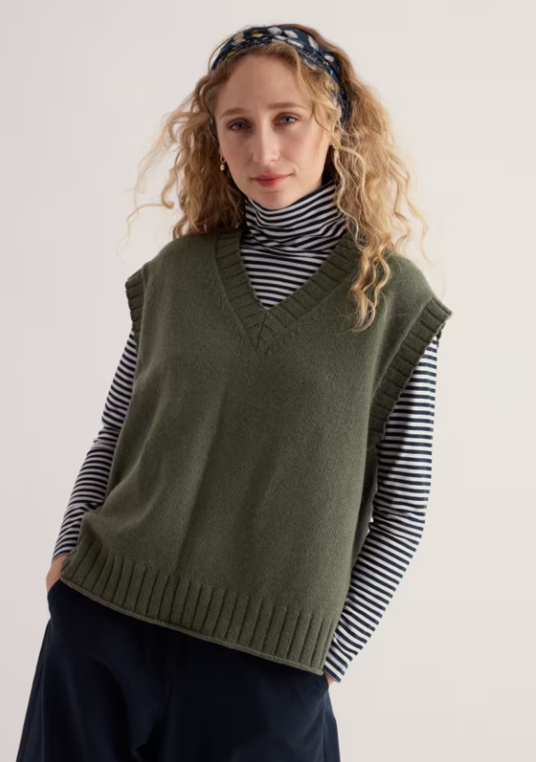 A woman wearing an olive green knit sweater vest over a black and white striped turtleneck, paired with dark navy pants and a patterned headband, photographed against a plain light background.