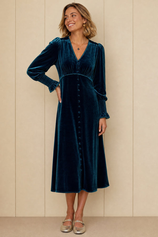 A woman wearing a long-sleeved dark teal velvet midi dress with a button-down front, standing against a beige paneled wall.