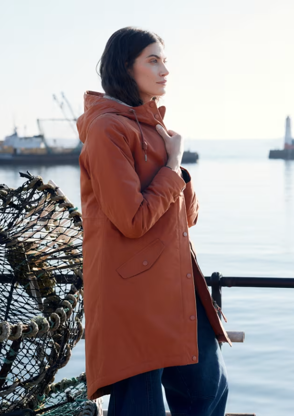A woman wearing a rust-orange hooded parka coat stands by the water, with fishing nets and boats visible in the background.