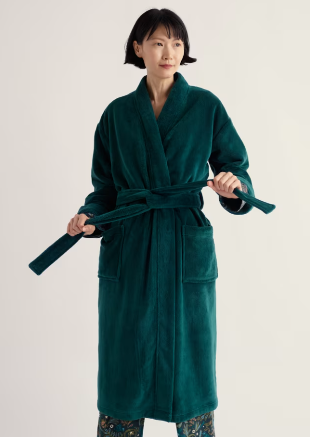 A woman wearing a plush long green bathrobe with a matching tie belt, standing against a plain light background.