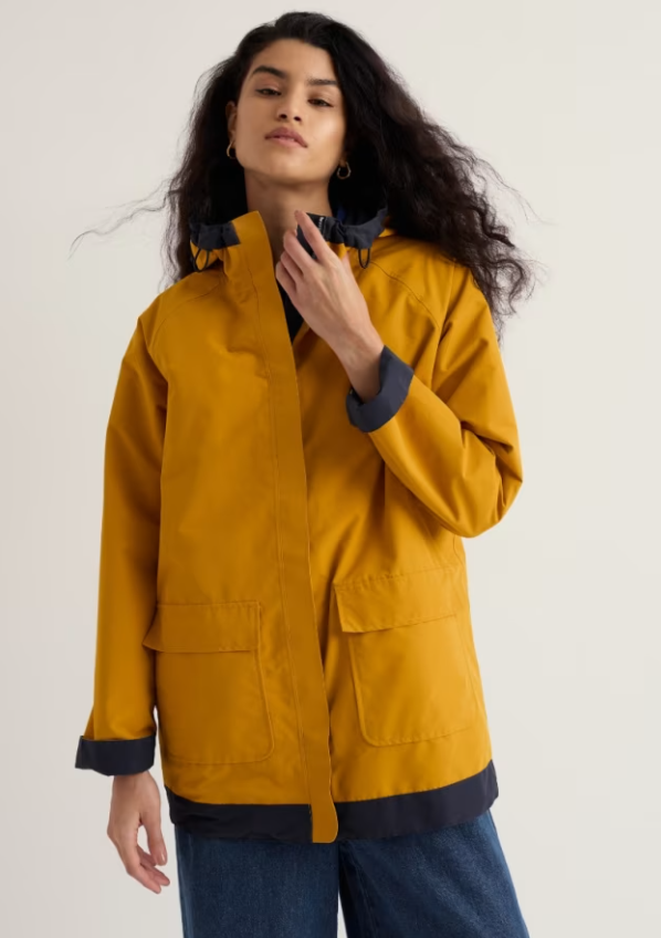 A woman wearing a mustard yellow hooded jacket with dark navy trim and cuffs, paired with blue jeans, photographed against a plain light background.