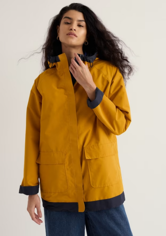 A woman wearing a mustard yellow hooded jacket with dark navy trim and cuffs, paired with blue jeans, photographed against a plain light background.