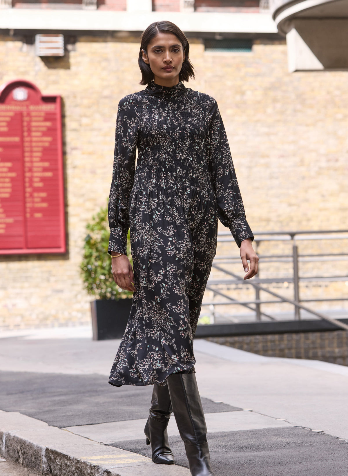 A woman walking outdoors on a paved path wearing a long-sleeved black midi dress with a white floral pattern, paired with black leather boots.