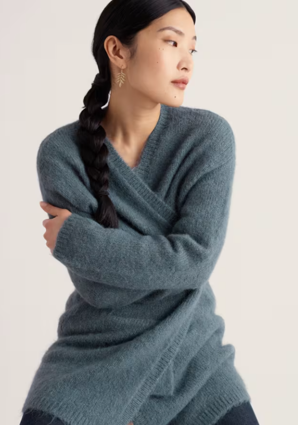 A woman wearing a soft, fuzzy knit V-neck cardigan in a muted blue-grey colour, posed against a plain off-white studio background.