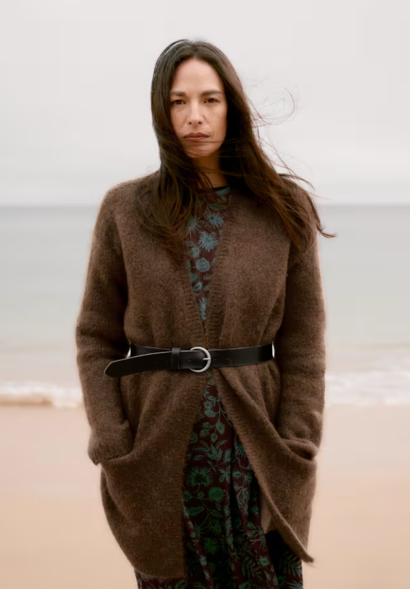 A woman stands on a beach wearing a brown open-front knit cardigan over a dark floral dress, cinched at the waist with a black belt.