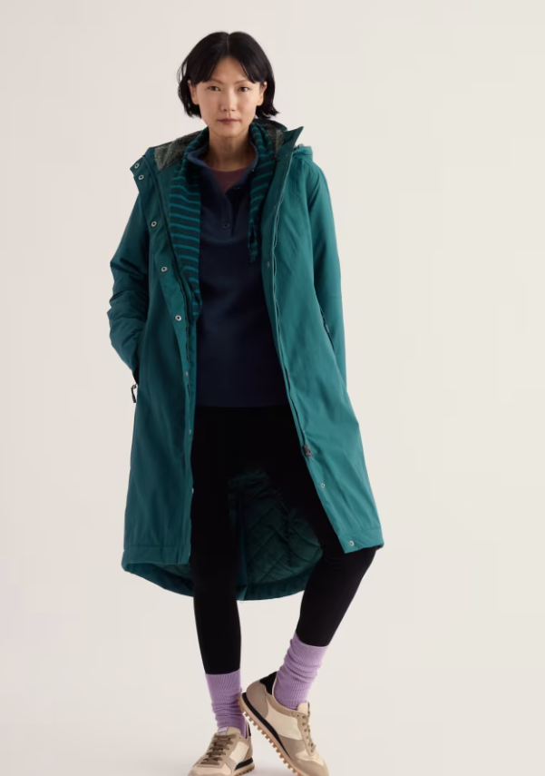 A model stands against a plain light background wearing a long teal hooded jacket over a dark striped top, black leggings, purple socks, and beige sneakers.