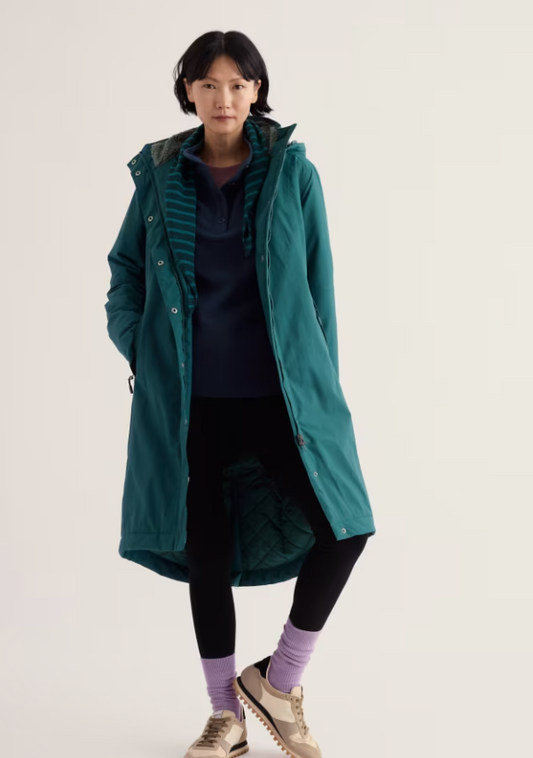 A model stands against a plain light background wearing a long teal hooded jacket over a dark striped top, black leggings, purple socks, and beige sneakers.