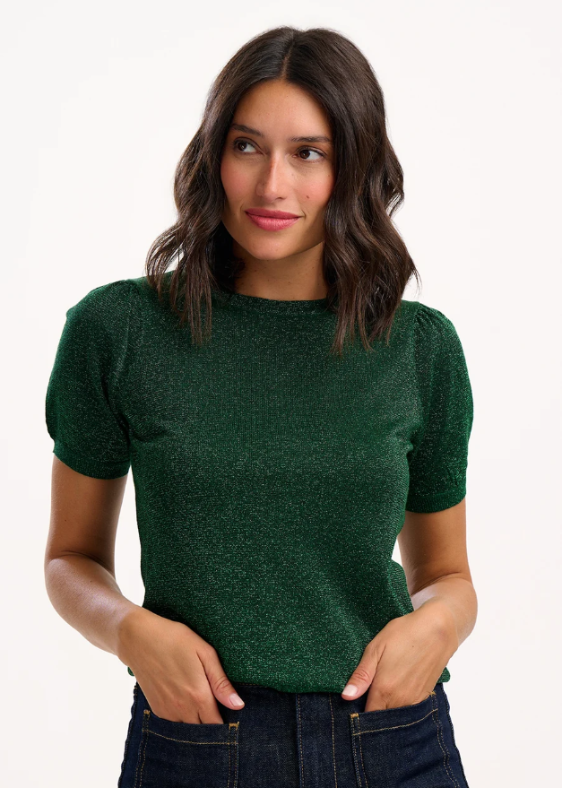 A woman wearing a dark green short-sleeve sparkle knit top with textured puff sleeves, paired with dark blue jeans, against a plain white studio background.