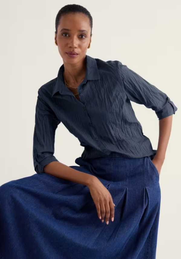 A woman seated against a plain light background wearing a dark blue crinkled button-up shirt and a matching dark blue skirt.