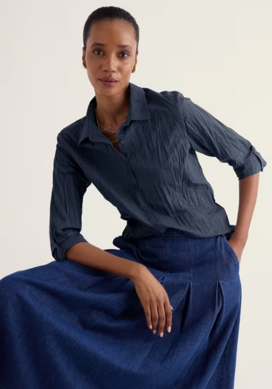 A woman seated against a plain light background wearing a dark blue crinkled button-up shirt and a matching dark blue skirt.