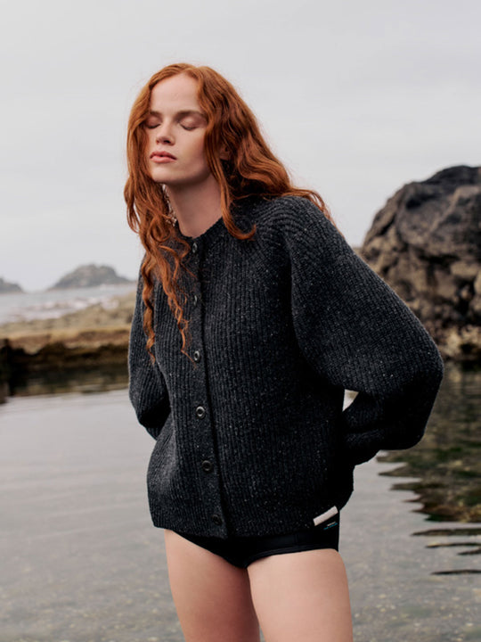 A woman with long red hair stands at a rocky coastal tide pool wearing a dark charcoal gray ribbed knit cardigan and black shorts.