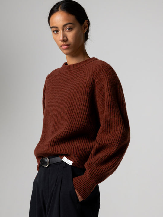 A woman wearing a rust-brown ribbed knit sweater and dark trousers, photographed against a plain light gray studio background.