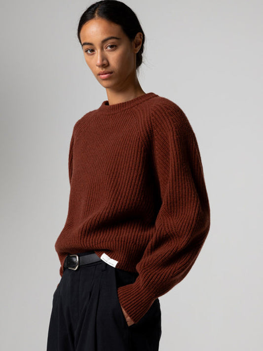 A woman wearing a rust-brown ribbed knit sweater and dark trousers, photographed against a plain light gray studio background.