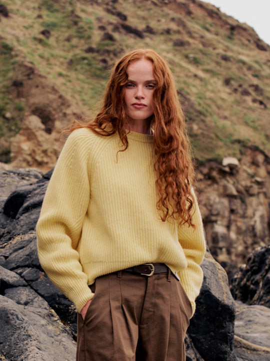 A woman with long red hair stands outdoors on rocky ground wearing a pale yellow ribbed knit sweater and brown trousers, with a grassy cliff in the background.