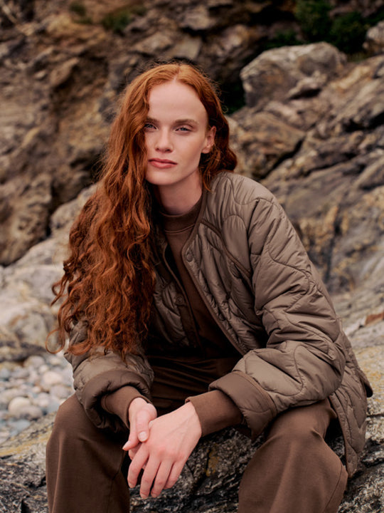 A woman with long, wavy red hair sits on a rocky outdoor surface wearing a quilted taupe-brown puffer jacket over a matching brown top and brown pants.