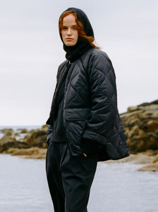 A model wearing a black quilted puffer jacket, black hooded balaclava, and black trousers, standing on a rocky shoreline under an overcast sky.