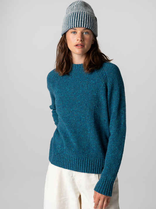 A woman wearing a teal blue heathered knit sweater, a blue and white striped beanie, and off-white trousers, photographed against a neutral light gray studio background.