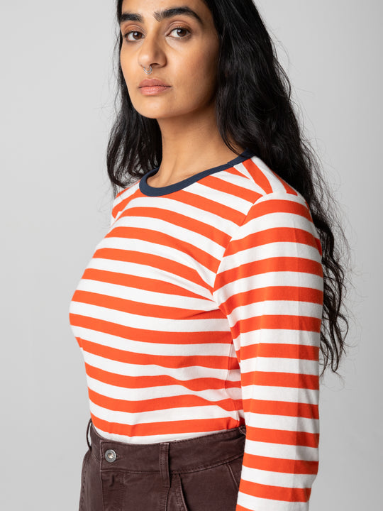 A woman wearing a long-sleeve crewneck top with horizontal orange and white stripes and a dark navy collar, paired with brown pants, against a solid light gray background.