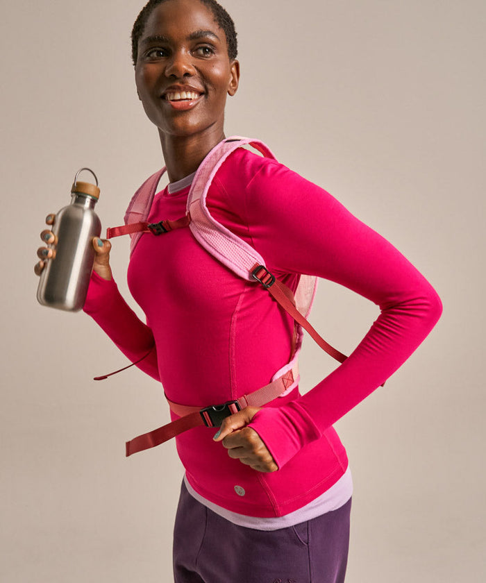 A woman wearing a bright pink long-sleeve athletic top and a pink backpack with red straps, holding a stainless steel water bottle against a plain beige studio background.
