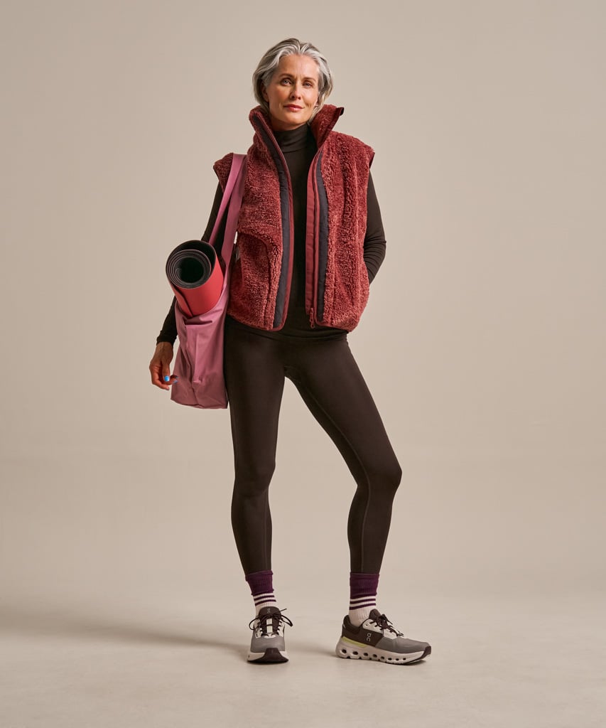A woman stands in a studio wearing a sleeveless textured red fleece vest over a black top, black leggings, and gray running shoes, while carrying a pink bag with a rolled pink mat.