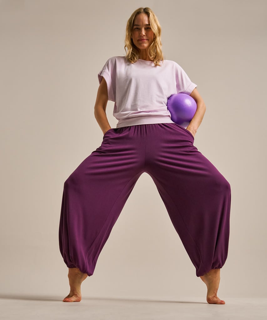 A woman modeling loose, baggy harem-style pants in deep purple, paired with a light lavender t-shirt and holding a purple exercise ball, against a solid light beige studio background.