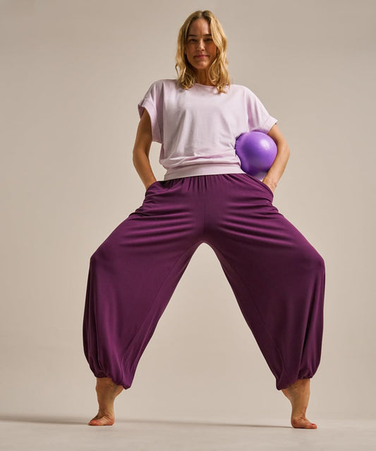 A woman modeling loose, baggy harem-style pants in deep purple, paired with a light lavender t-shirt and holding a purple exercise ball, against a solid light beige studio background.