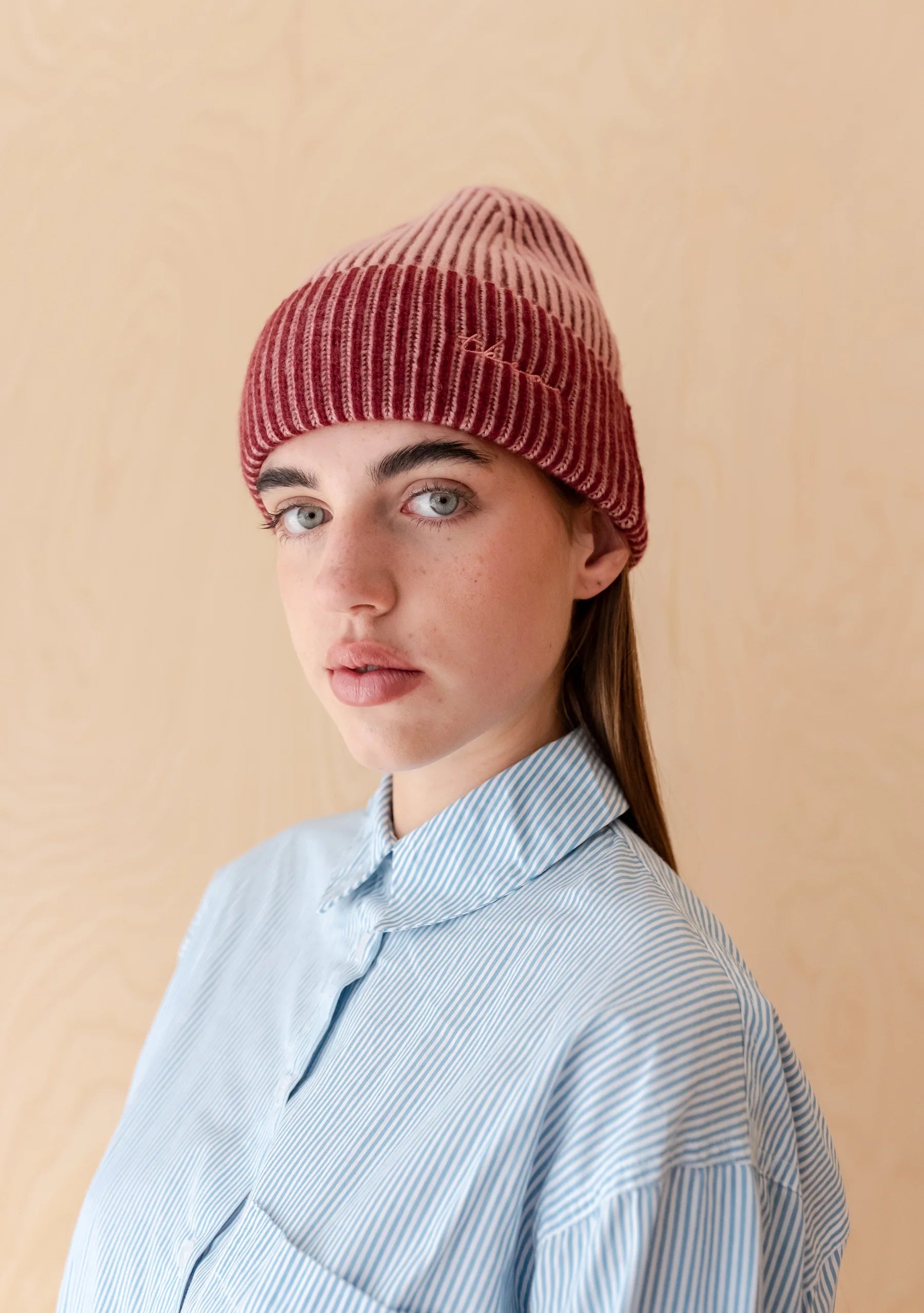 Person wearing a red and pink knitted beanie and light blue shirt against a beige background. Hat is by TBCo and filtered for the Soft Summer and Soft Autumn colour seasons by Shop My Palette.