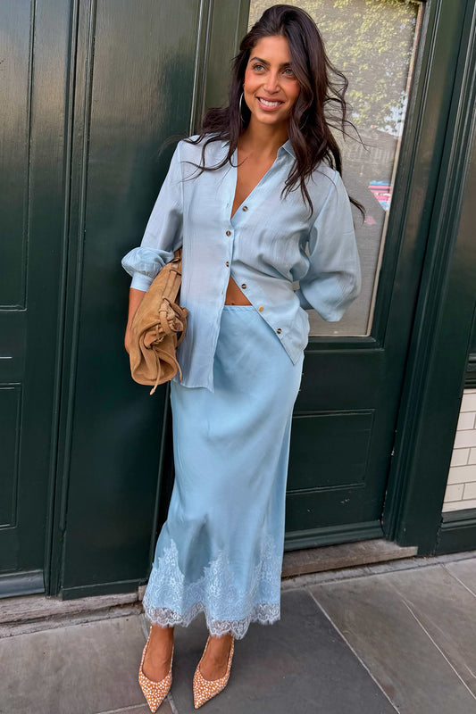 A woman stands outdoors in front of a dark green door wearing a light blue satin button-up shirt and matching midi skirt with lace trim, paired with tan pointed heels and a tan clutch bag.