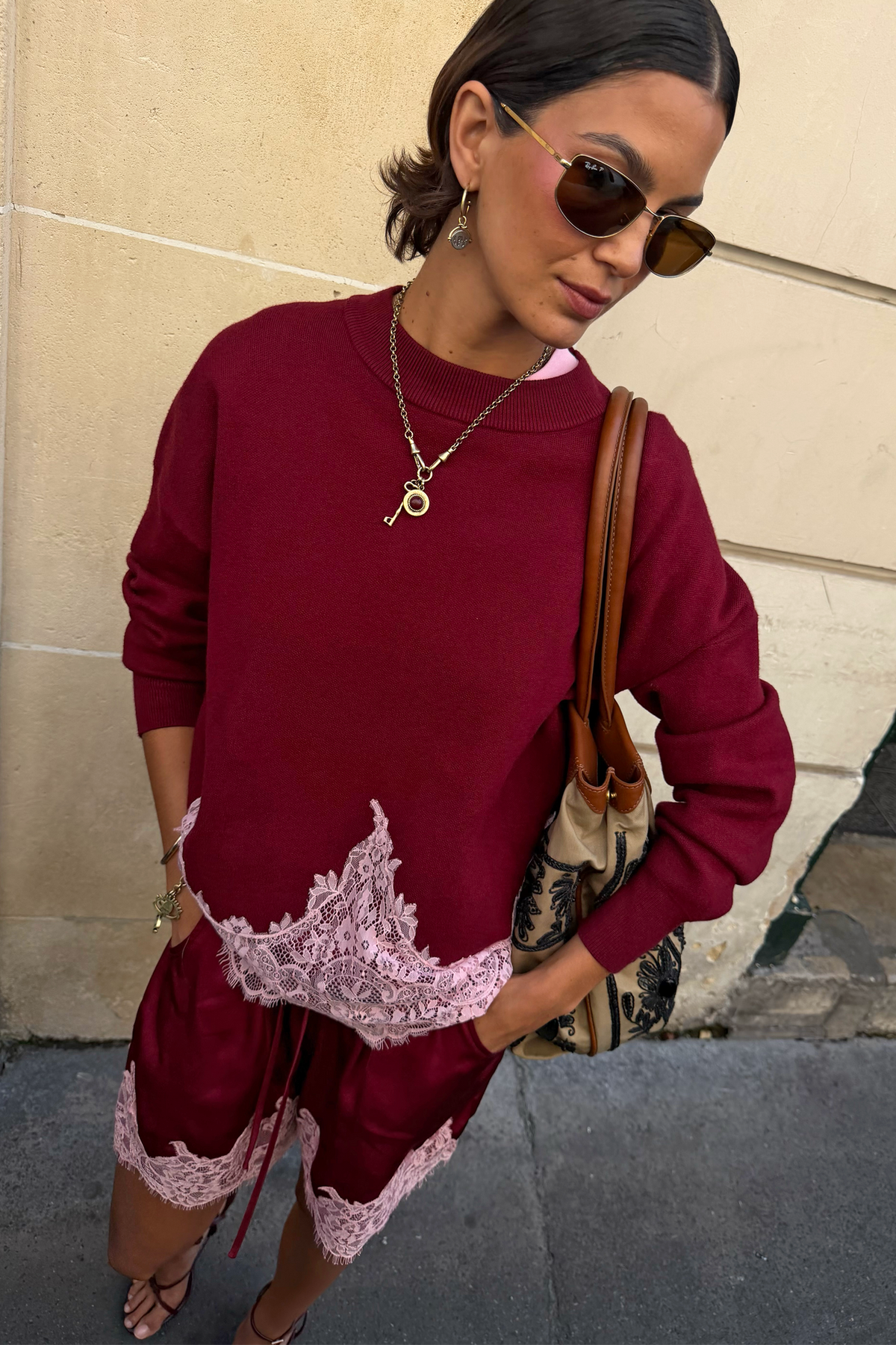 A woman wearing a burgundy sweatshirt and matching shorts set with light pink lace trim, styled with sunglasses, a key necklace, and a brown shoulder bag against a beige wall.