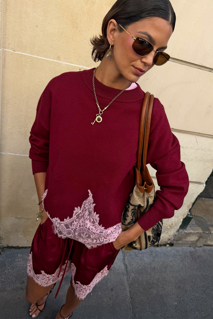 A woman wearing a burgundy sweatshirt and matching shorts set with light pink lace trim, styled with sunglasses, a key necklace, and a brown shoulder bag against a beige wall.