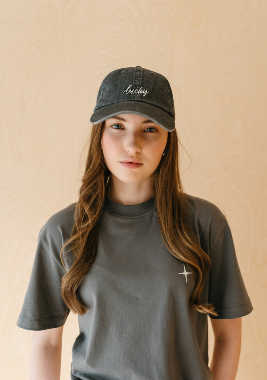 Washed charcoal grey cotton baseball cap with curved brim and white embroidered “lucky” lettering on the front