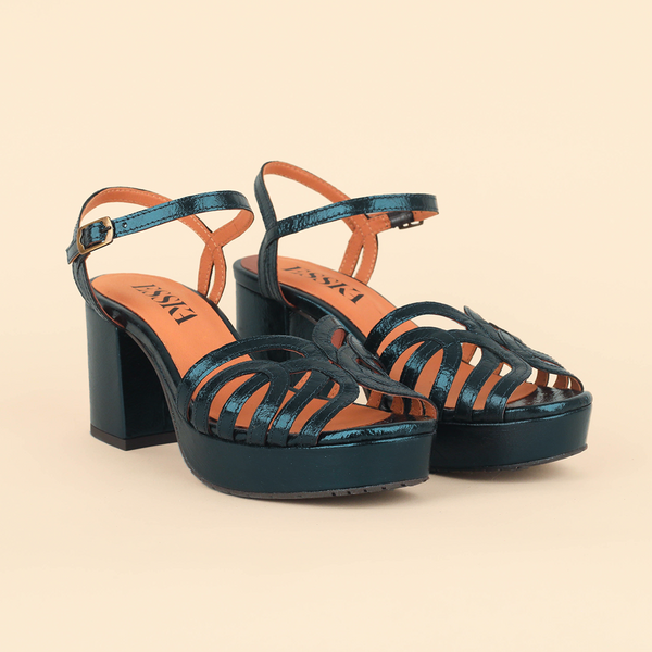 A pair of metallic teal platform sandals with cutout straps, ankle straps, and block heels, shown on a solid beige background.