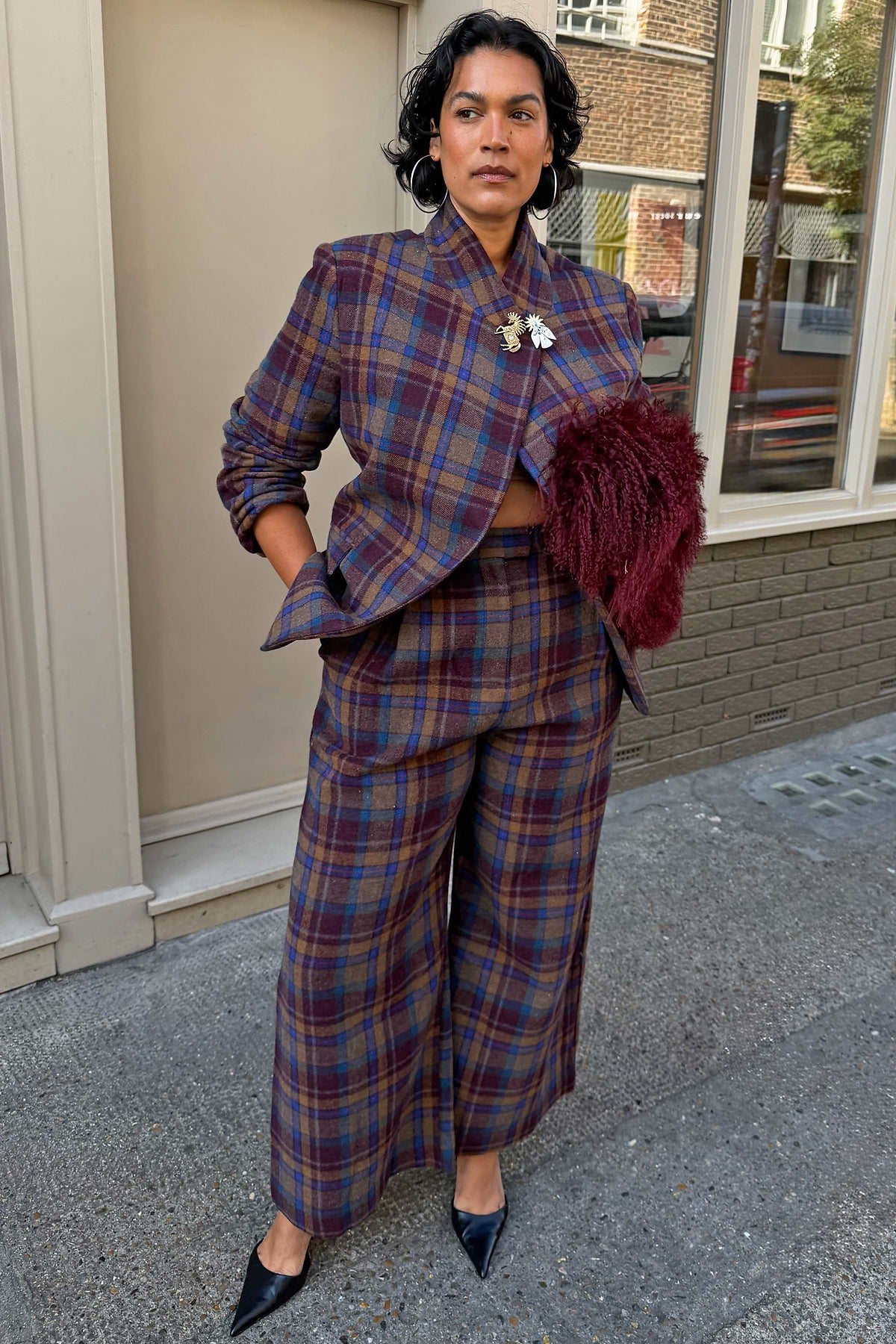 A model stands on a city pavement wearing a matching plaid blazer and wide-leg trousers set in shades of purple, blue, and red, paired with a dark burgundy textured clutch and black pointed heels.