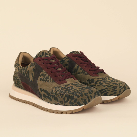 A pair of low-top trainers with an olive green and black abstract patterned upper, burgundy laces, and a thick cream midsole with a gum outsole, shown on a solid beige background.