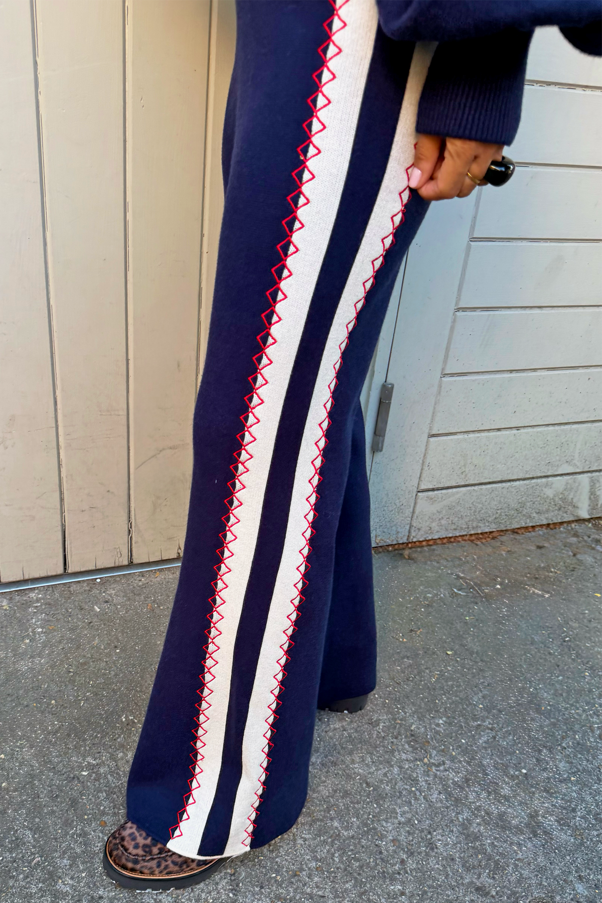 A close-up of a person wearing navy blue wide-leg knit trousers with a bold white side stripe bordered by red zigzag stitching, standing outdoors against a beige and grey panelled wall.
