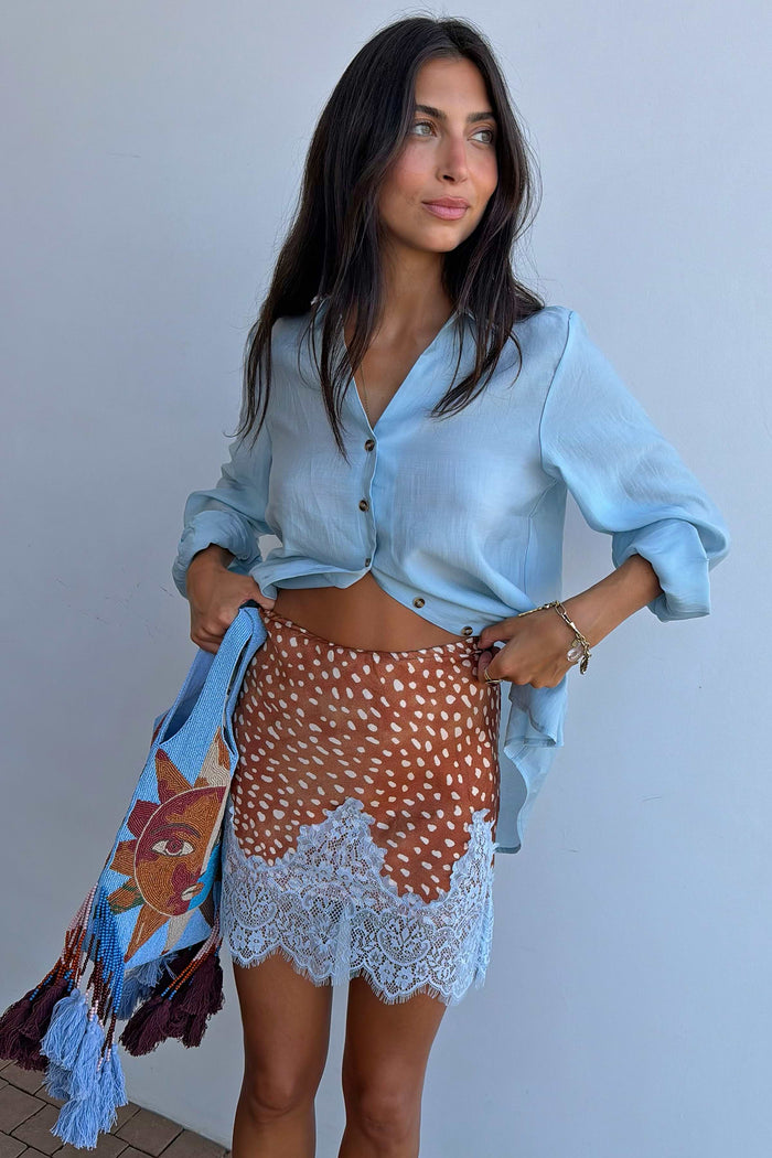 A woman modeling a light blue button-up shirt and a brown patterned skirt with white lace trim, holding a blue patterned scarf against a plain light gray wall.