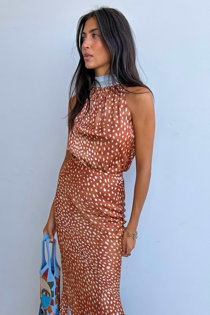 A woman wearing a sleeveless caramel top and skirt with a white spotted pattern, standing against a light blue wall and holding a blue patterned tote bag.