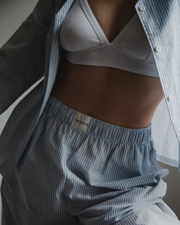 Close-up of a person wearing ribbed light grey bra and matching striped pyjama set with a visible NUDEA waistband label, layered under an open striped button-up shirt.