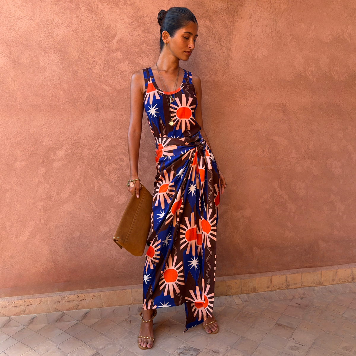 A woman stands against a textured terracotta wall wearing a sleeveless wrap-style maxi dress with a bold sunburst print in navy, rust orange, and cream. She is holding a brown leather clutch and wearing brown strappy heels.