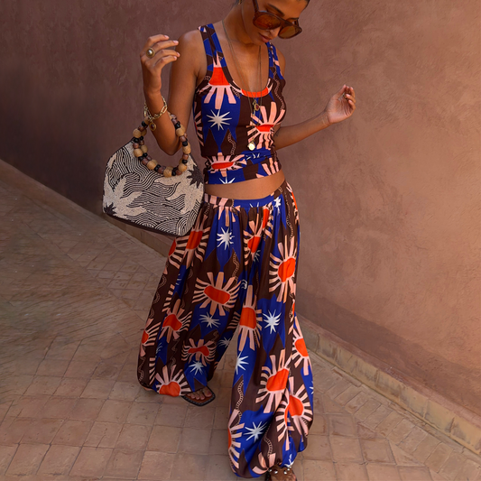 A woman wearing a matching two-piece set with a colourful sunburst pattern in blue, brown, orange, and white, posing against a textured brown wall.