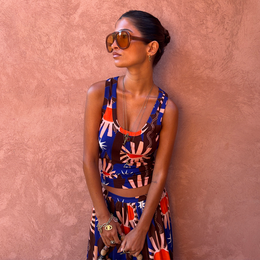 A woman wearing a matching sleeveless top and skirt set with a blue, orange, and brown abstract floral print, styled with large brown-tinted sunglasses, standing against a textured terracotta wall.