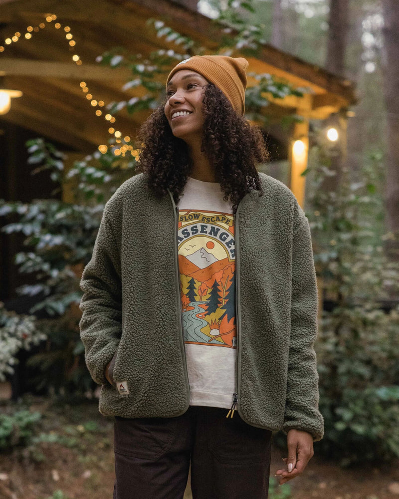 Woman wears a dusty olive sherpa fleece, PASSENGER t-shirt and orange beanie hat.