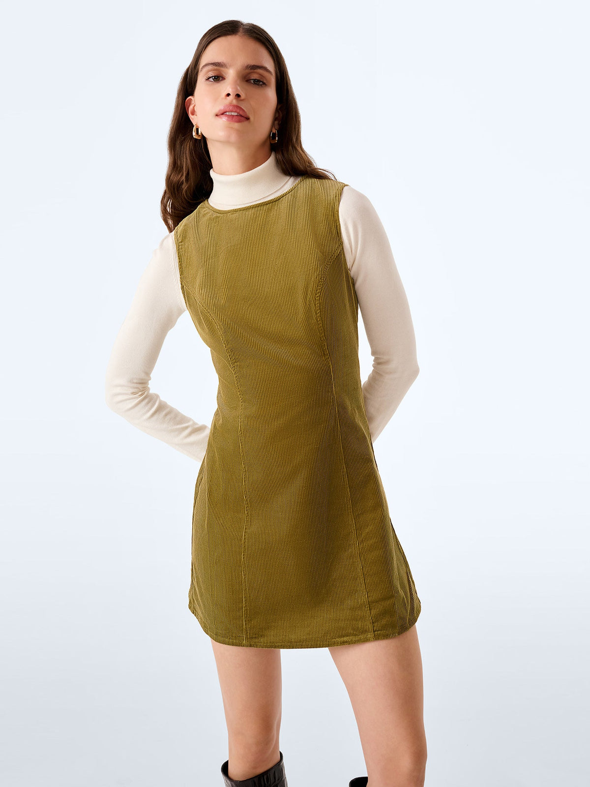 A woman modeling a sleeveless olive green corduroy mini dress over a cream long-sleeve turtleneck, set against a plain light gray studio background.