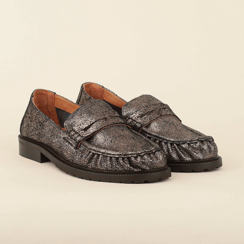 A pair of metallic silver moccasin-style loafers with a textured glitter finish, brown leather lining, and low block heels, photographed on a solid light beige background.
