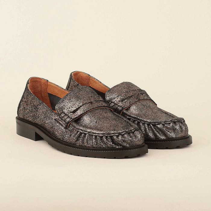 A pair of metallic silver moccasin-style loafers with a textured glitter finish, brown leather lining, and low block heels, photographed on a solid light beige background.